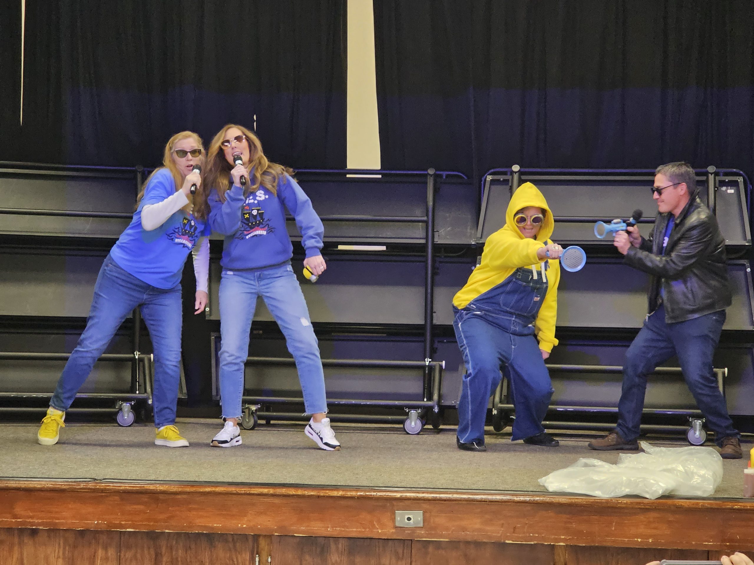 Staff members singing and dancing on school stage for a fundraising activity.